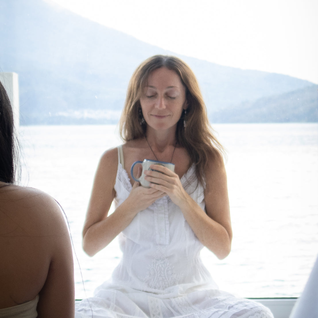 Alla, in a white sleeveless dress, sits by a large window overlooking Mount Fuego and Lake Atitlán, holding a mug of Keith's ceremonial cacao drink with both hands in reverence. Another person is partially visible in the foreground.