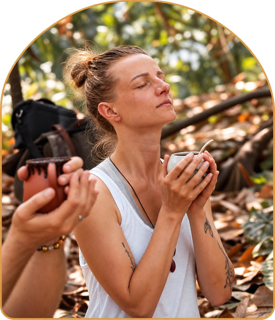 Samantha, in a white tank top, stands in prayer the cacao forest in Guatemala at Homecoming, holding a white ceramic mug filled with warm Keith's Ceremonial Grade cacao, while another person on the left holds a terracotta mug. You can see dry leaves and trees in the background.