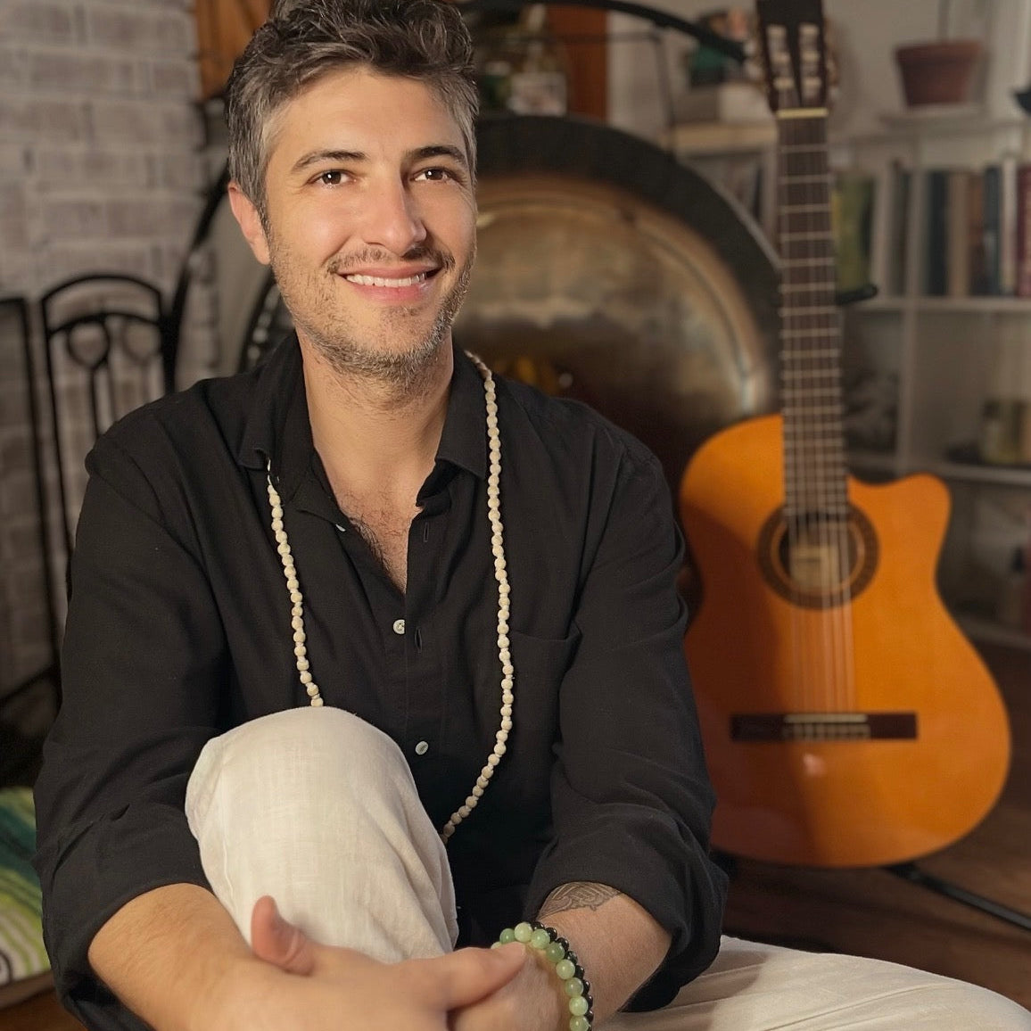 Keith's Cacao practitioner Avi Burnbaum sits cross-legged indoors, wearing a black button-up shirt, white pants, a long white beaded necklace, and a green beaded bracelet. An acoustic guitar and a large metal gong are visible in the background of the room.