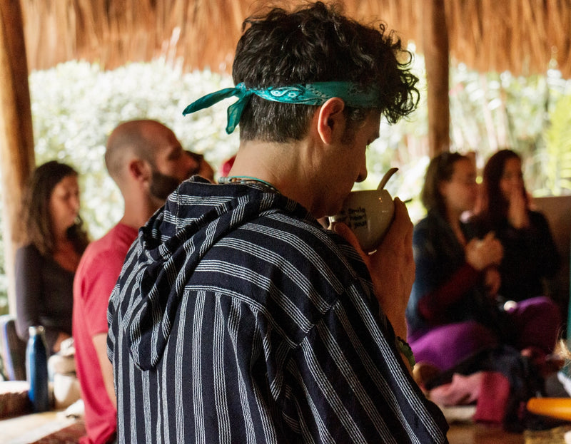 Avi in a striped hoodie and teal bandana drinking ceremonial cacao from a mug inside the Palapa,  a thatched-roof outdoor setting, in La Paz during Keith's Cacao Homecoming with other people blurred in the background.