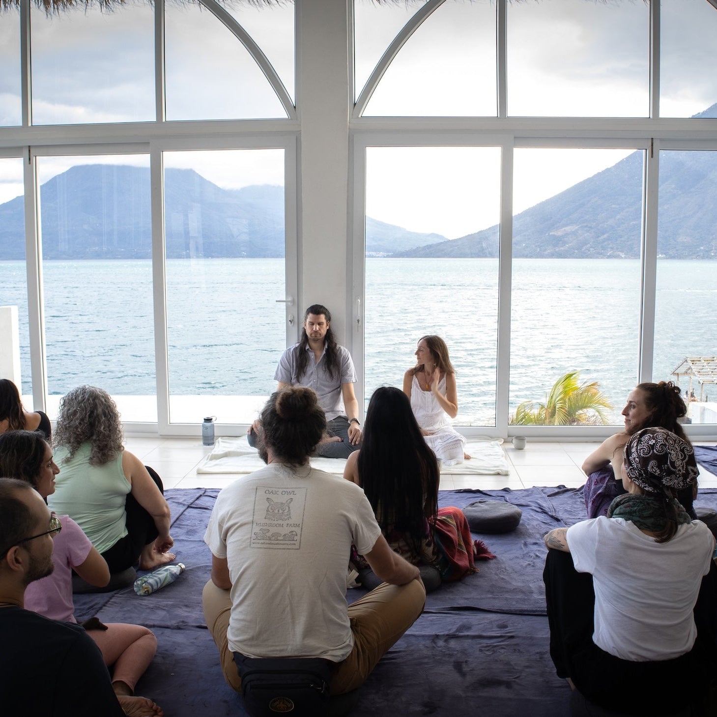 Group of people sitting on the floor in a room with large windows overlooking a scenic view.