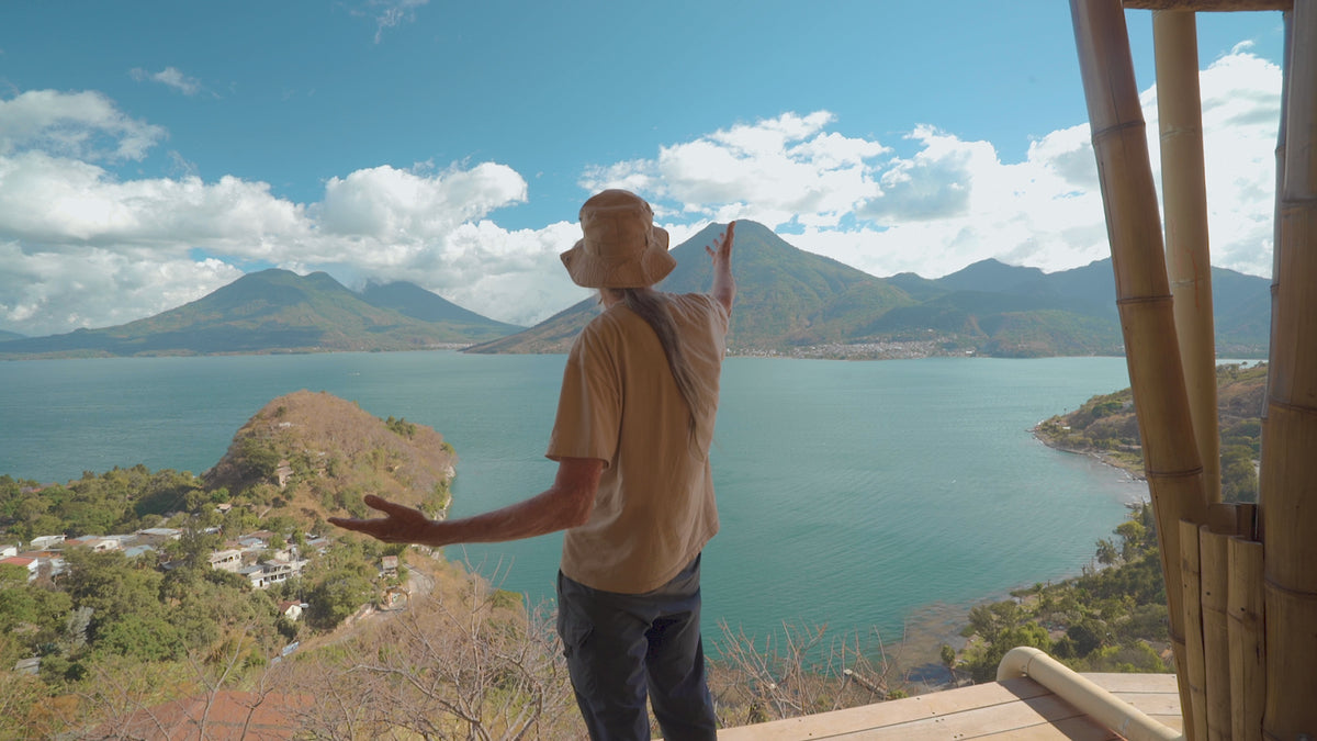 Keith Wilson, wearing a beige T-shirt, blue jeans, and a beige bucket hat, stands on the wooden balcony at Eagle's Nest in Guatemala, overlooking Lake Atitlán and Mount Fuego in the background, blue water, and a partly cloudy sky, gesturing towards the landscape.