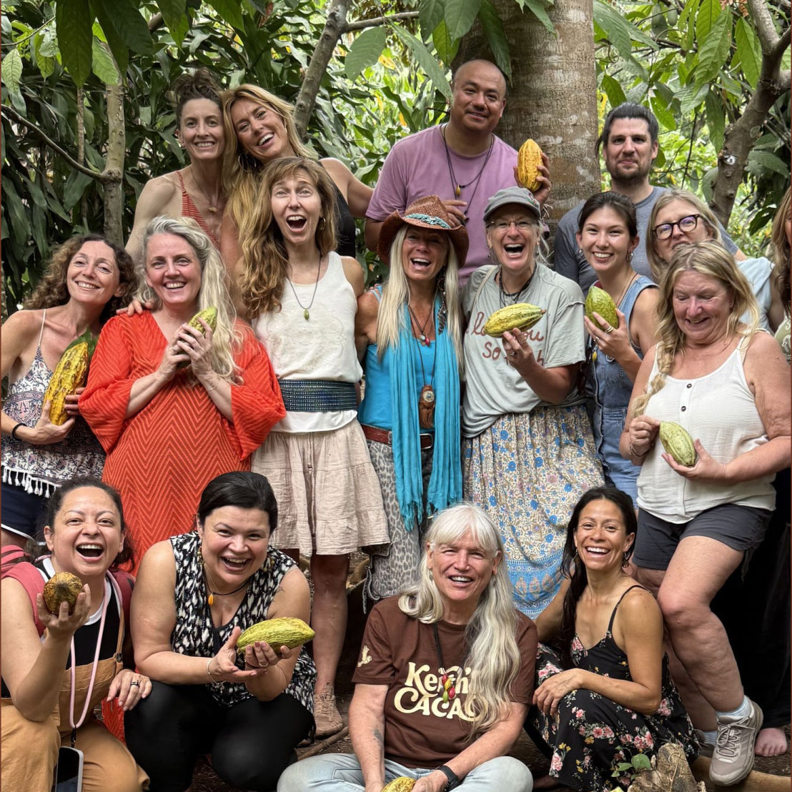 Keith's Cacao Homecoming 2026 participants are posing together in the cacao forest, each holding a yellow cacao pod. One person in the front row wears a brown t-shirt with 'Keith's Cacao' printed on it.