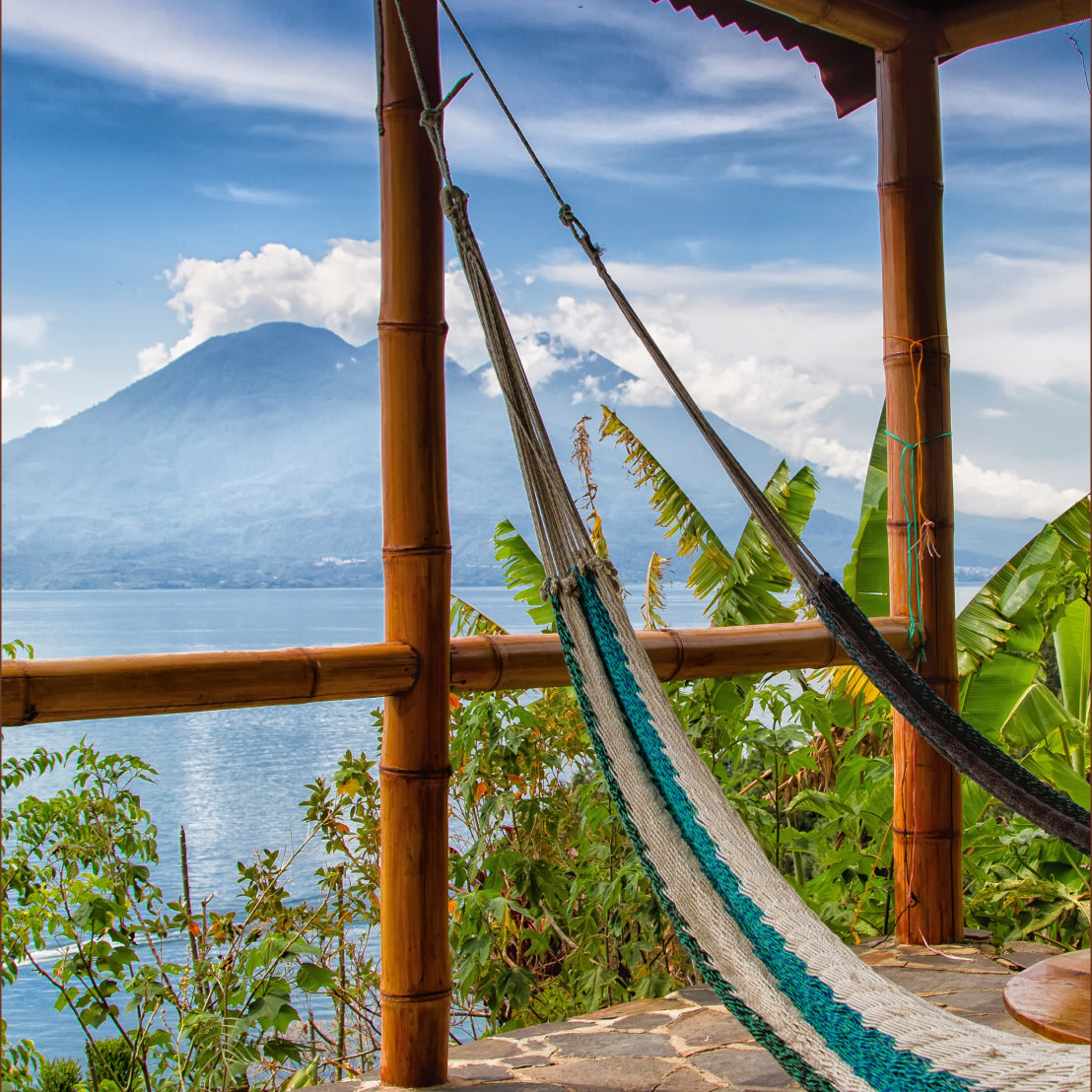 A woven hammock with teal and cream stripes hangs between bamboo posts on a wooden deck, overlooking the scenic Lake Atitlán with Mount Fuego in the background.