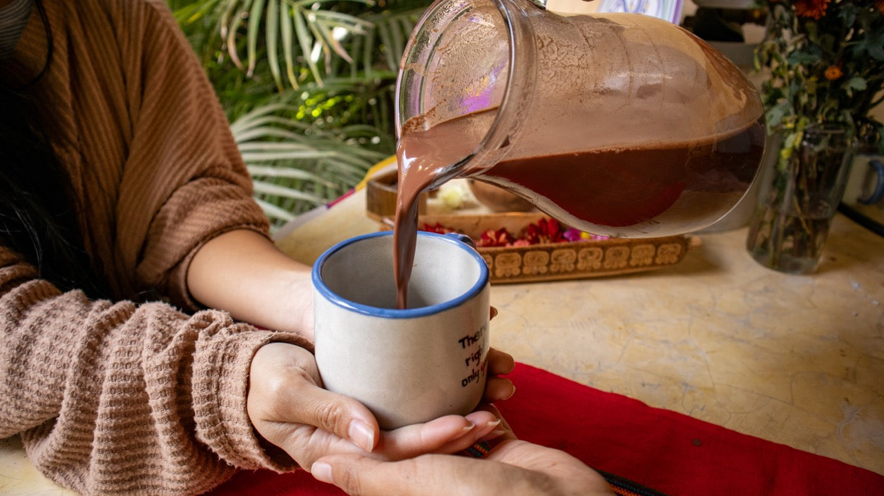 Person pouring cacao from a glass pitcher into a white mug with a blue rim.