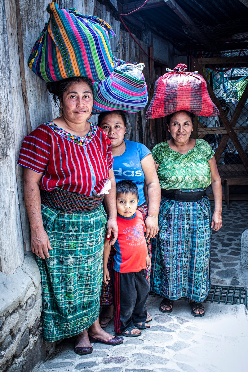 Keith's Cacao tostadoras and one of their children standing together, wearing traditional clothing and carrying cacao beans on their heads that they will take home to lightly toast and hand-peel..