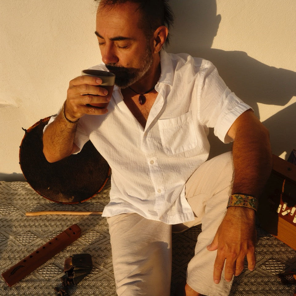 Keith's Cacao practitioner Carlos Marcus in a white short-sleeve button-up shirt and light pants sits on a patterned rug against a textured wall, holding a small cup of cacao to his lips. A drum and other objects are visible in the room